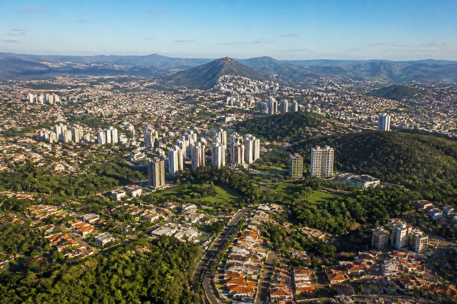 Vista aérea de várias cidades próximas a Belo Horizonte com prédios, áreas residenciais, parques verdes e colinas ao redor.
