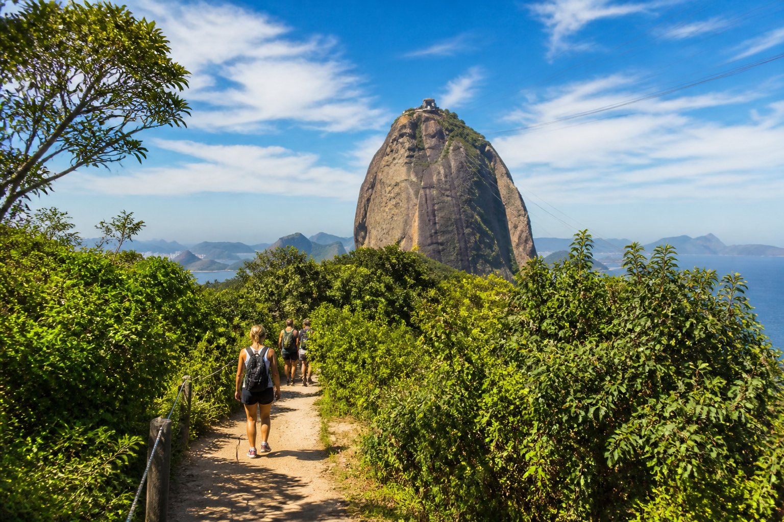 Trilha da Urca com caminho cercado por vegetação verde e o Pão de Açúcar ao fundo sob céu azul.